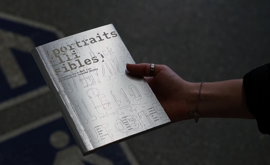 A person holds a small, silver/gray artist’s book titled “portraits illisibles.” The cover contains faint line drawings, diagrams, and fragmented typography. The person wears rings and a delicate bracelet, while the dark background and directional lighting emphasize the book and hand.