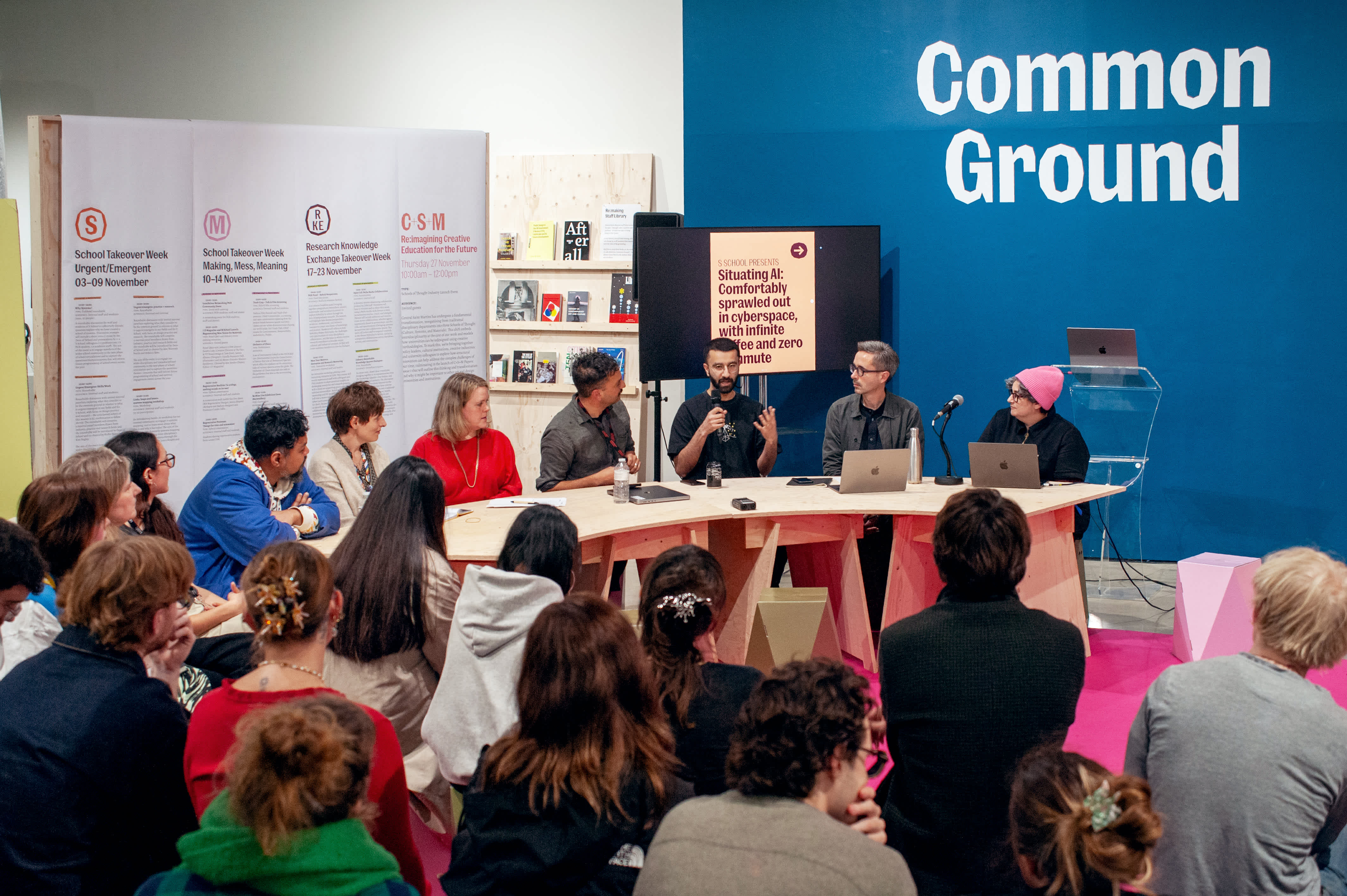 Panelists in discussion seated around a table under a sign in caps stating 'Common Ground'. An audience watches on