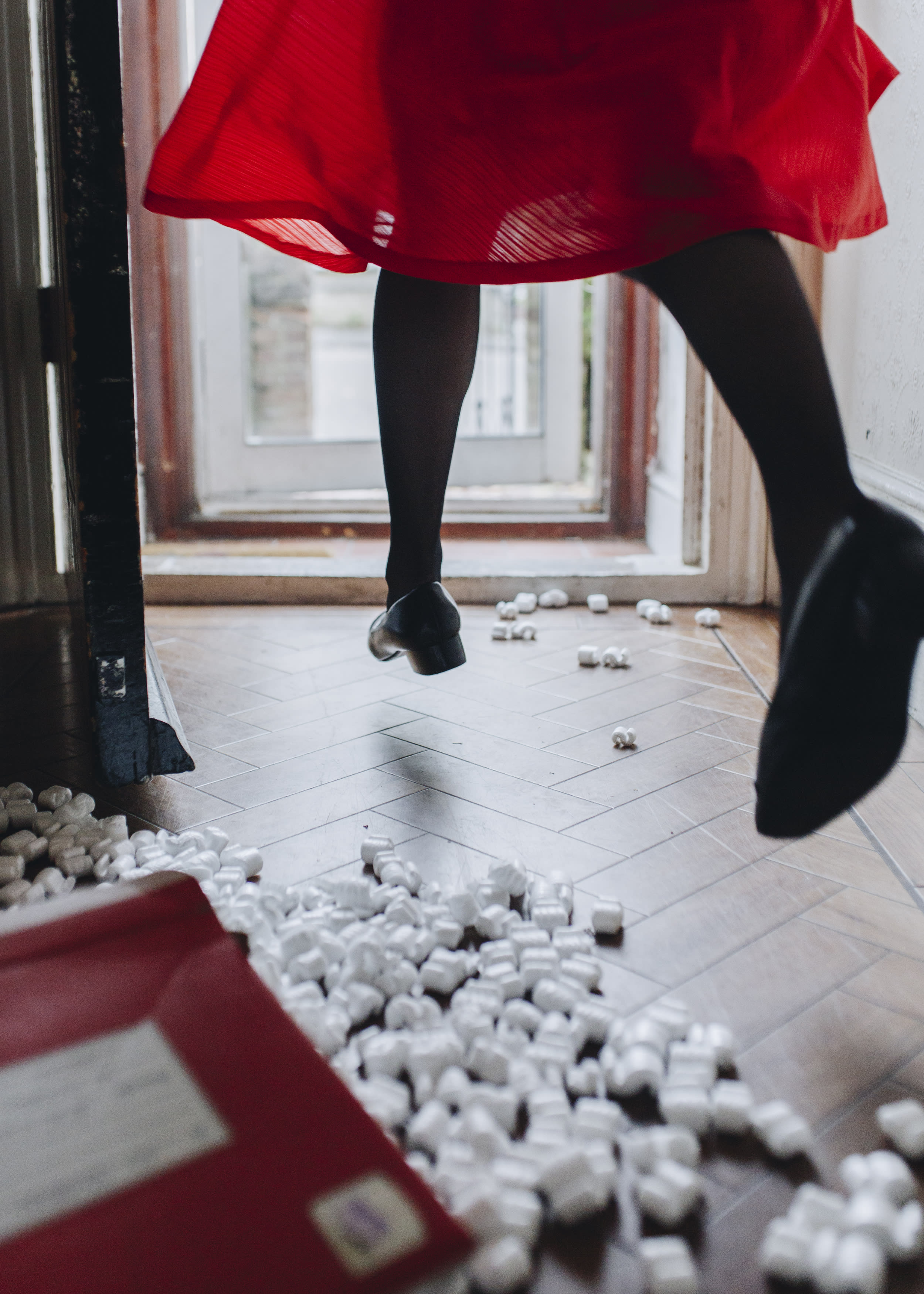 A person wearing a red dress, black stockings and heels, skipping towards a door, only their legs are visible.