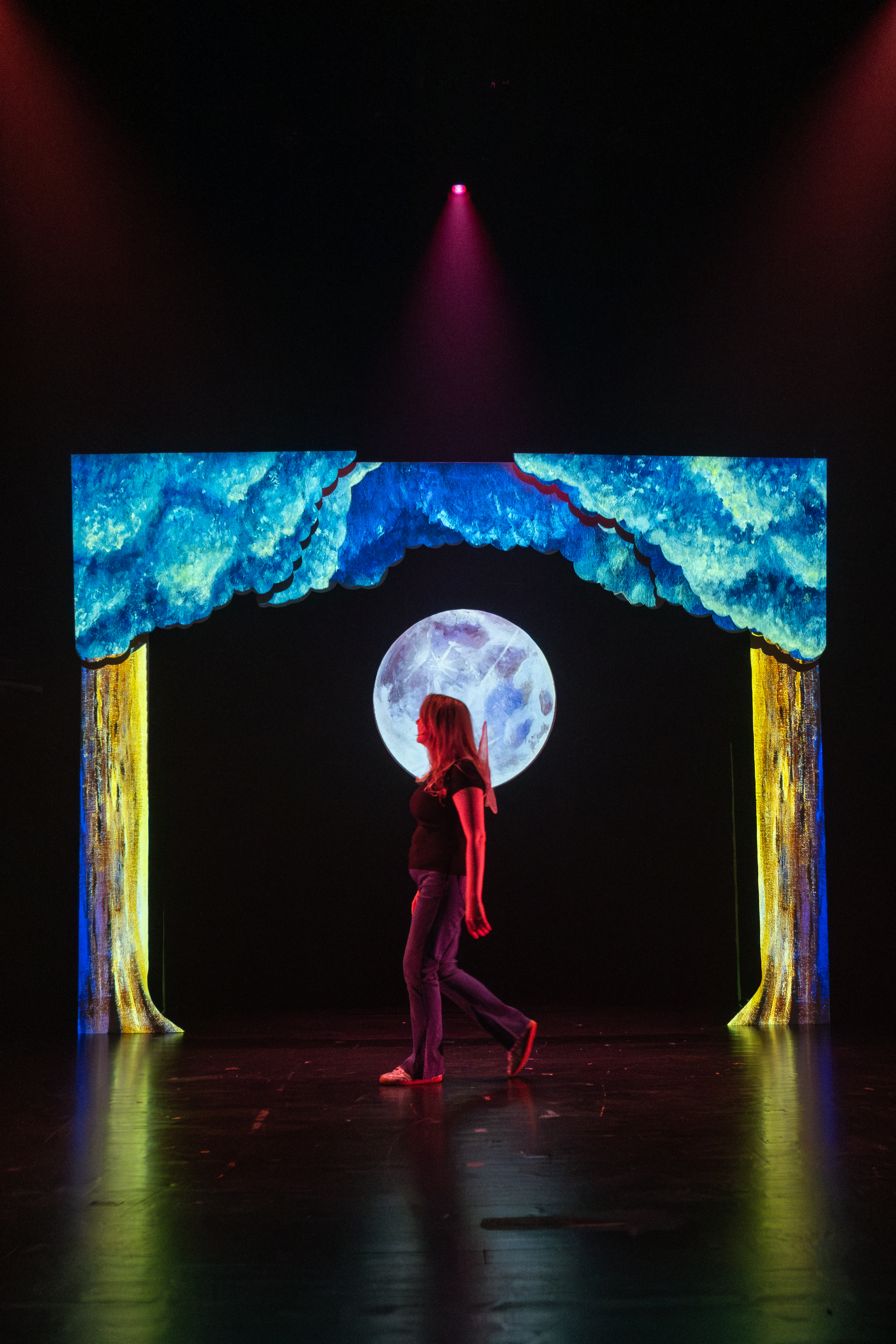 A girl looks shocked as she holds two styrofoam cups to her chest, she is performing ona set with orange and blue lighting