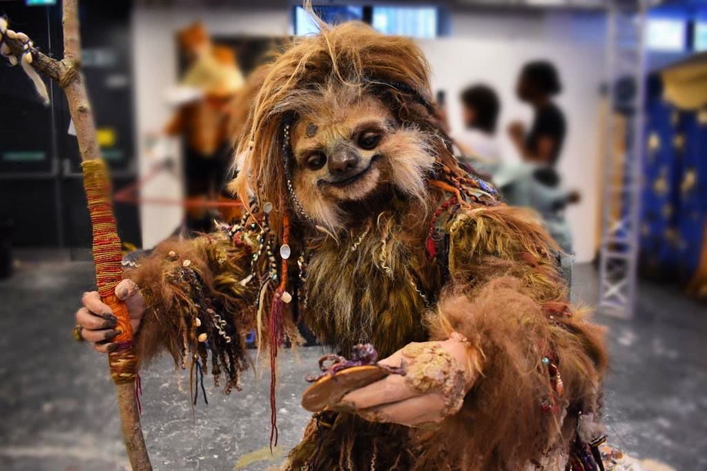 Person in an elaborate, furry sloth costume holding a staff and an object, standing indoors with blurred people in the background.