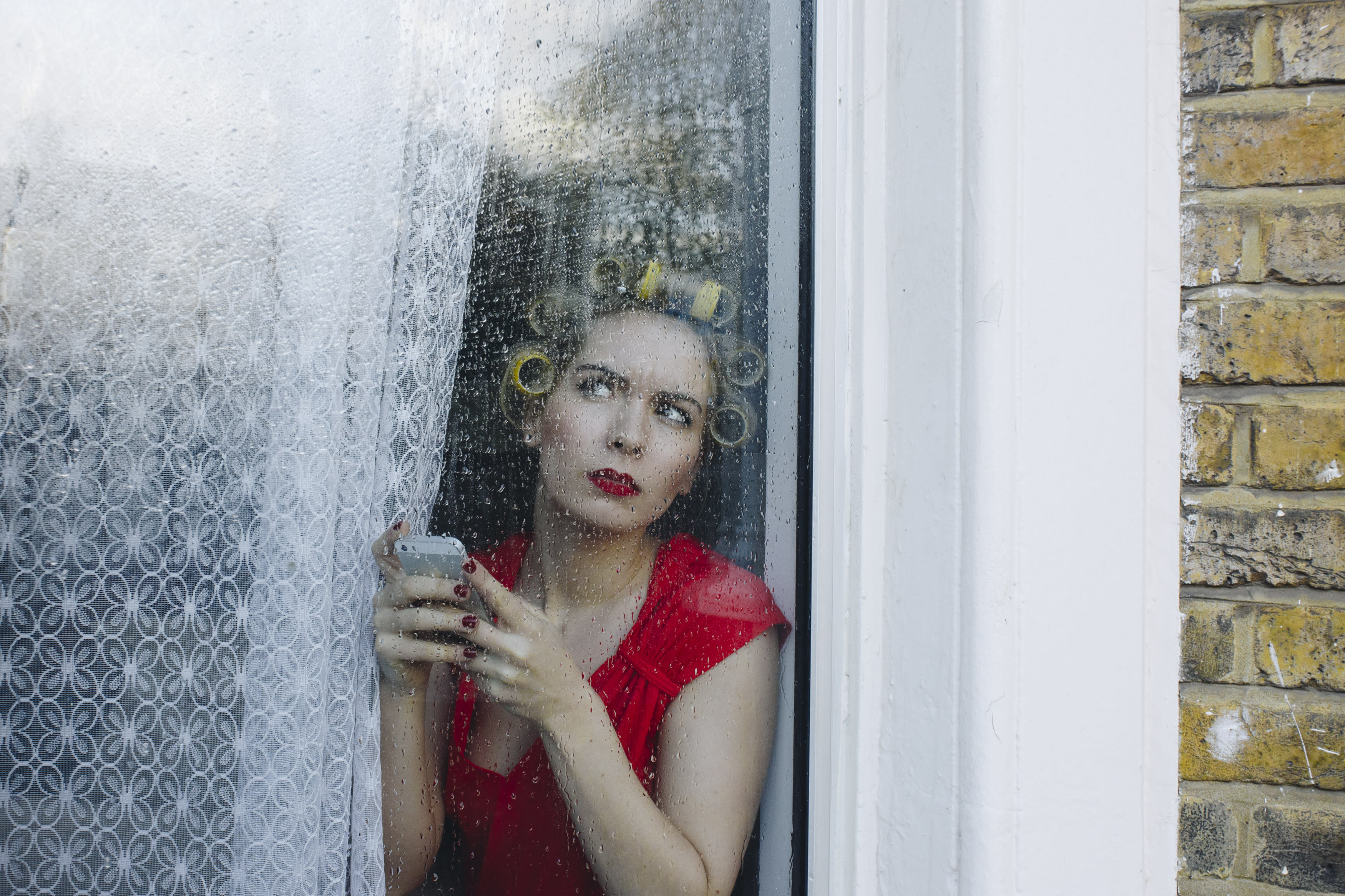 A person wearing a red dress and yellow rollers in their hair, looking out of their window with a phone in their hands.