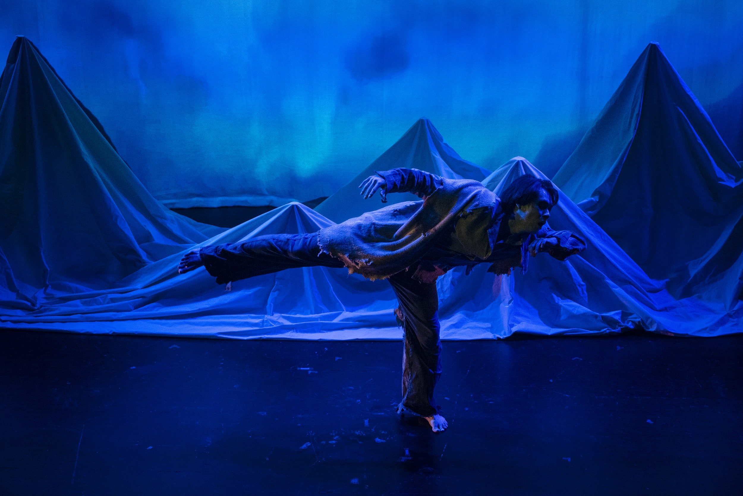 A girl walks in front of a set wearing wings, the set has a large moon and blue trees