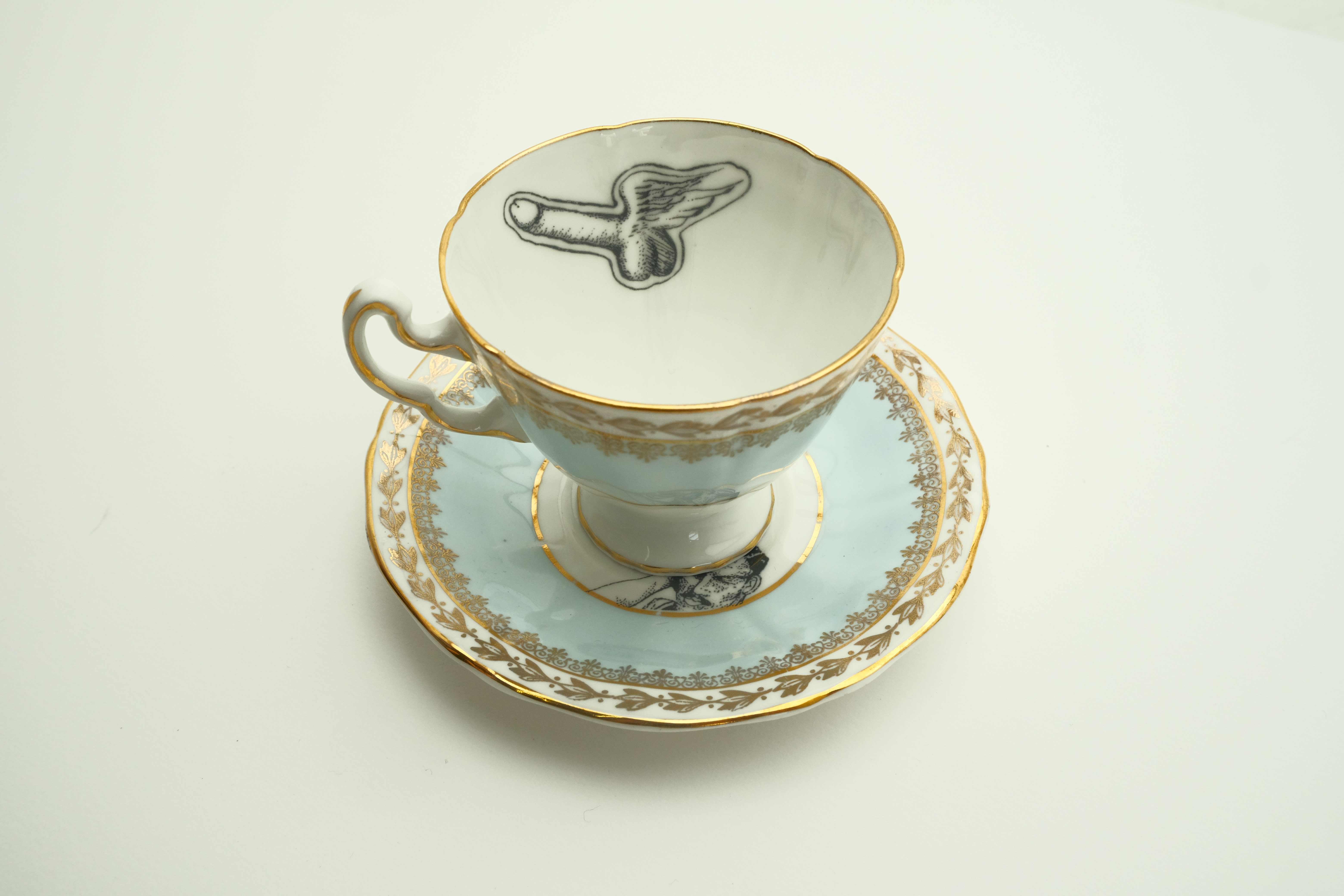 A porcelain teacup and saucer in pale blue and white with decorative gold trim. A design inside the cup is a floating penis with wings