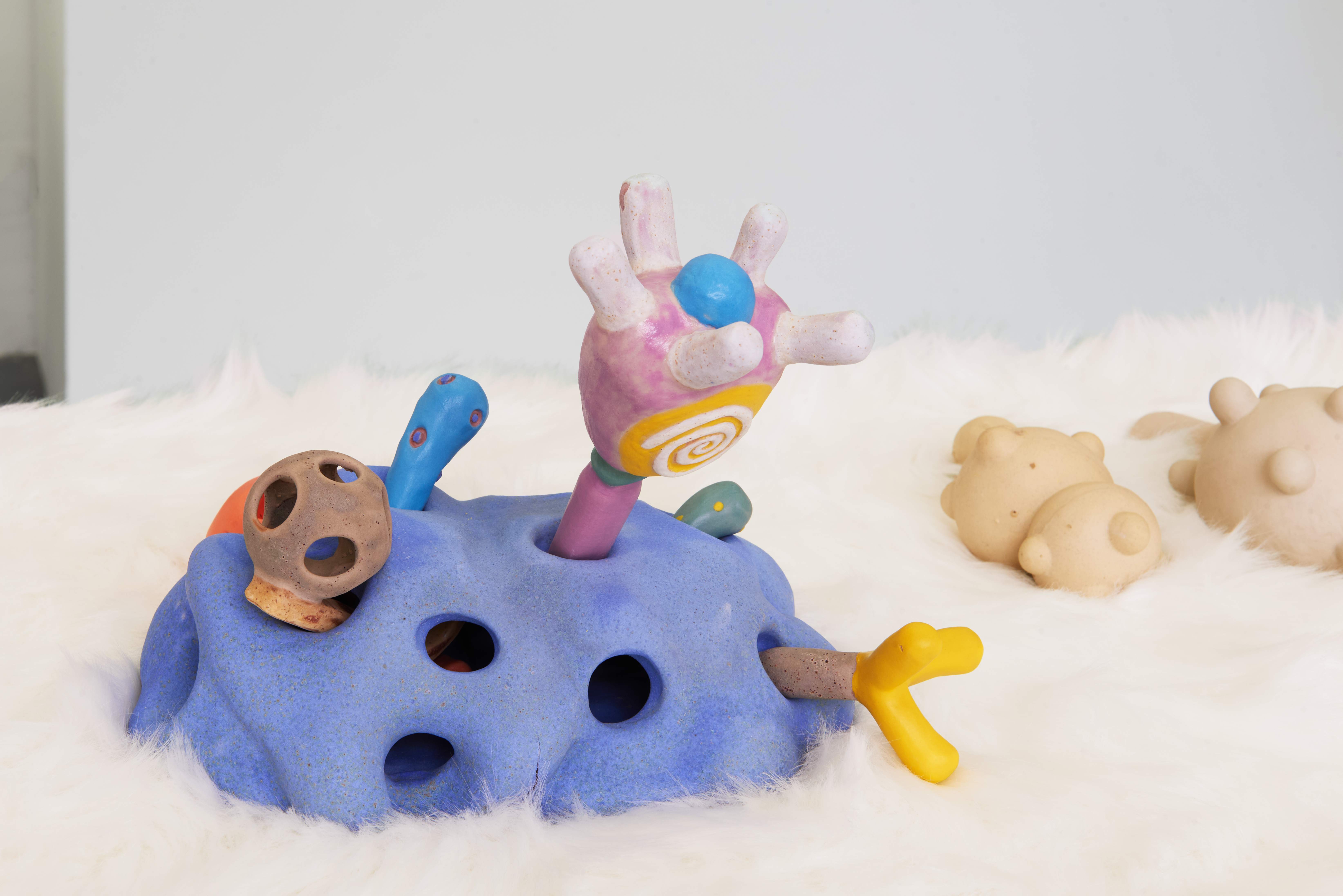 Colourful abstract ceramic sculpture with rounded forms and protruding shapes, displayed on a white furry surface alongside smaller cream ceramic objects.