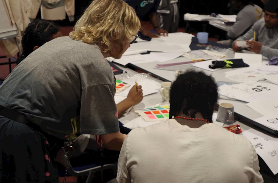 Participants are seated around a table working with pens, postits and paper. A tutor is leaning over and demonstrating to one of the participants.