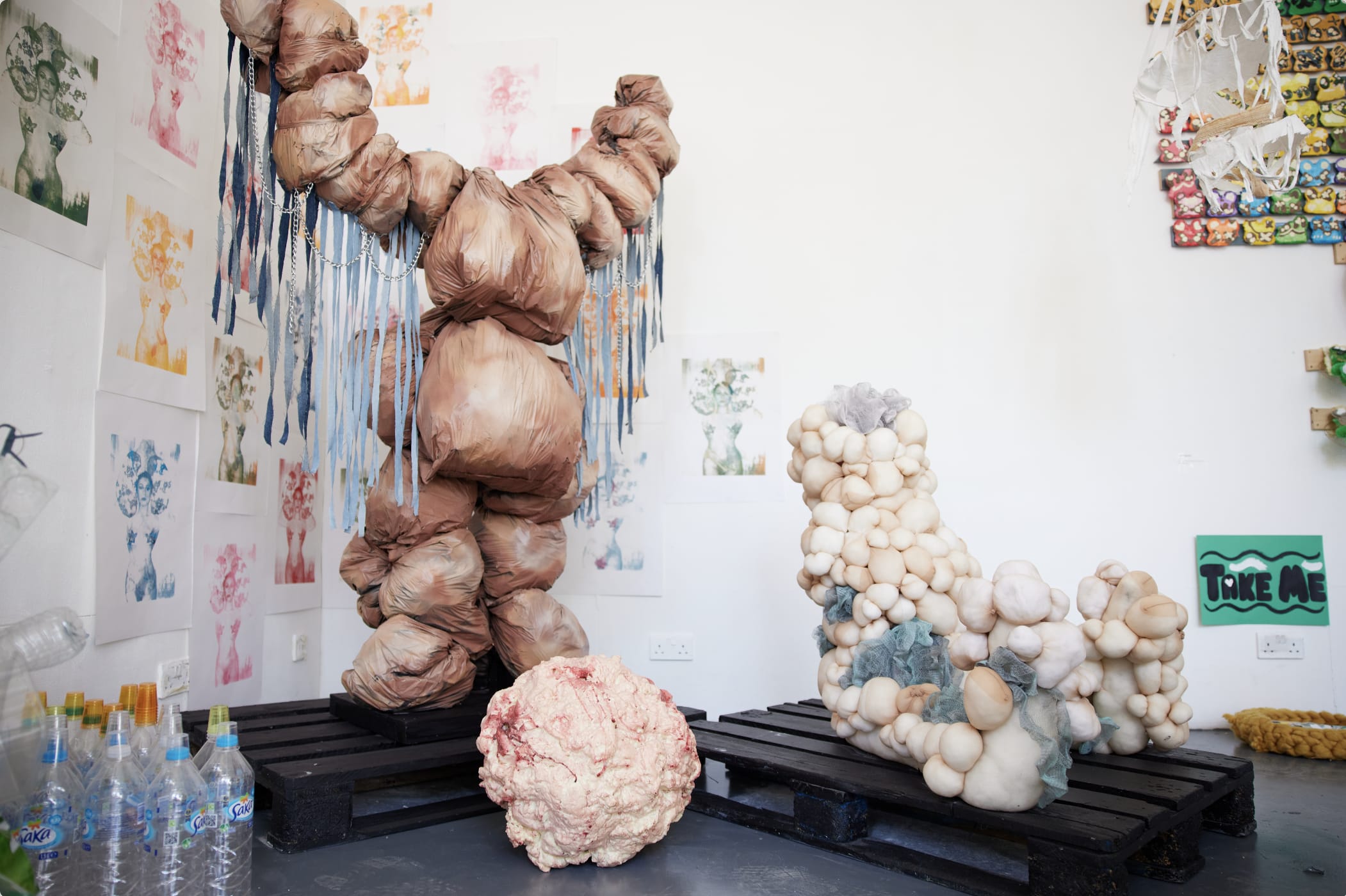 A large brown-beige sculpture representing a body made from rounded fabric compnents. The arms of the sculpture are raised upwards with denim and metal fringe hung down from the arms. 