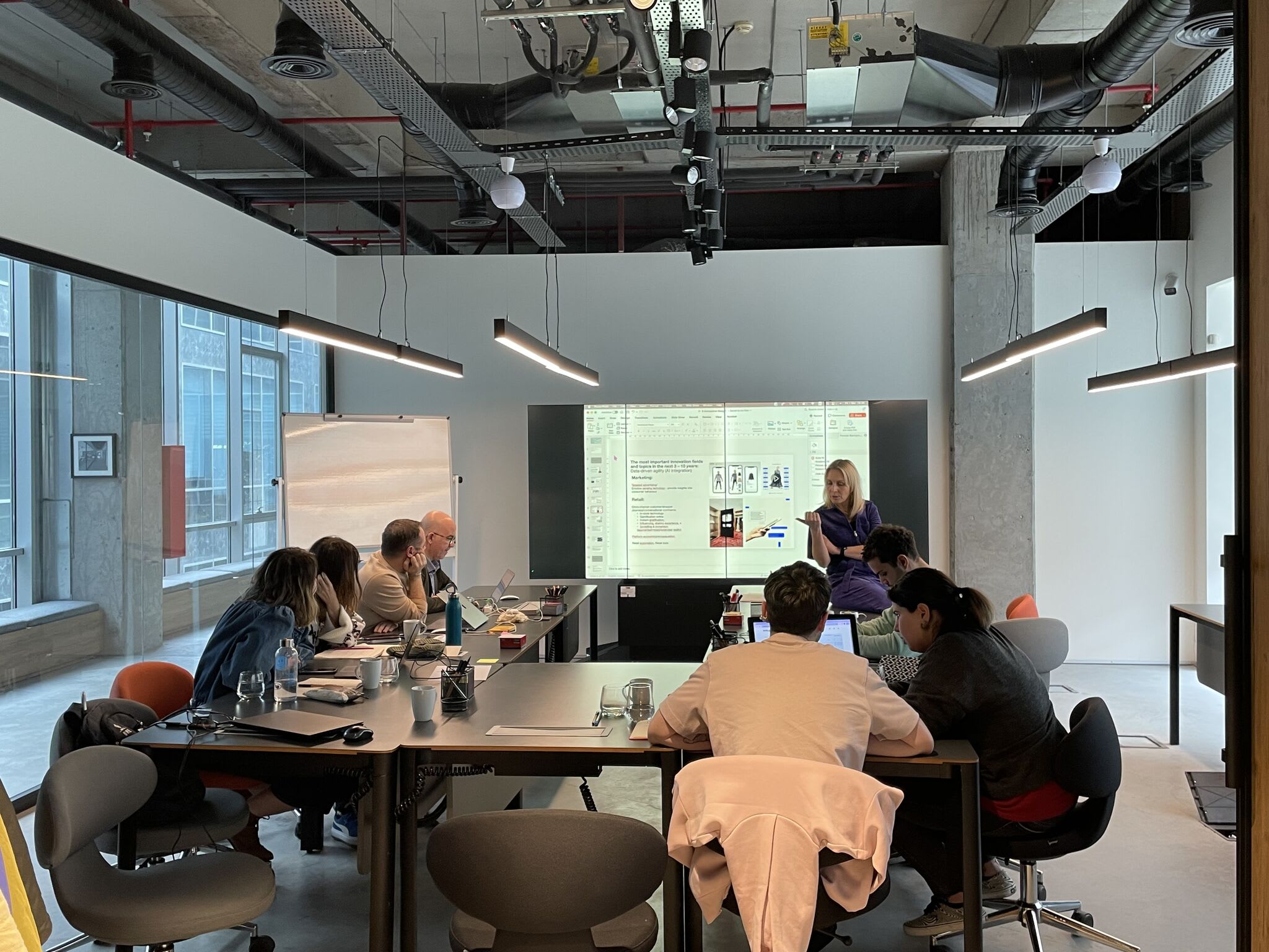 A group of individuals sit around a U shaped table facing towards a person standing up in front of a screen and providing training.
