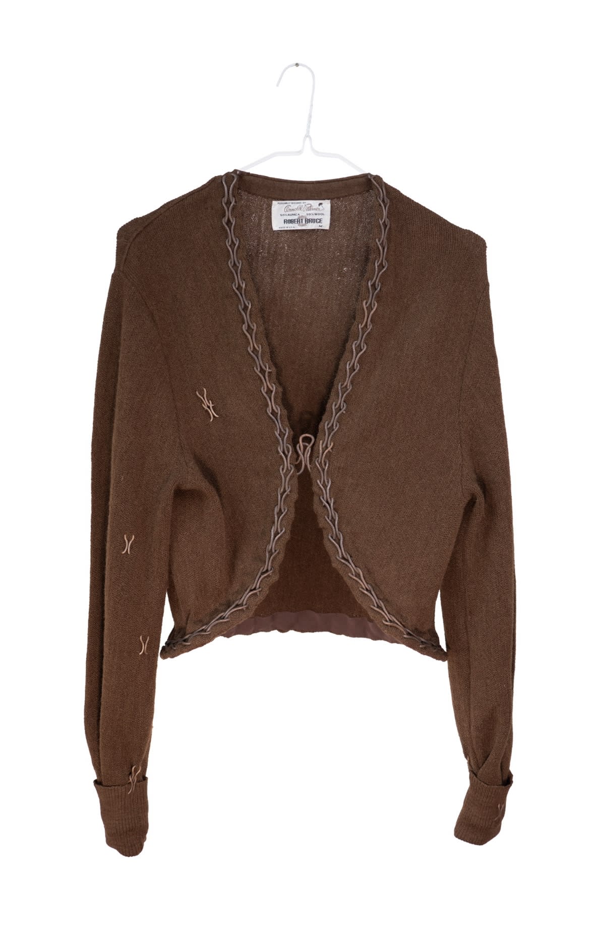 A brown cardigan has been repaired using recycled heirloom jewellery. There is visible decorative mending and a repeating symbol pattern as a trim and fastening. These appear to be bronze or rusted metal.