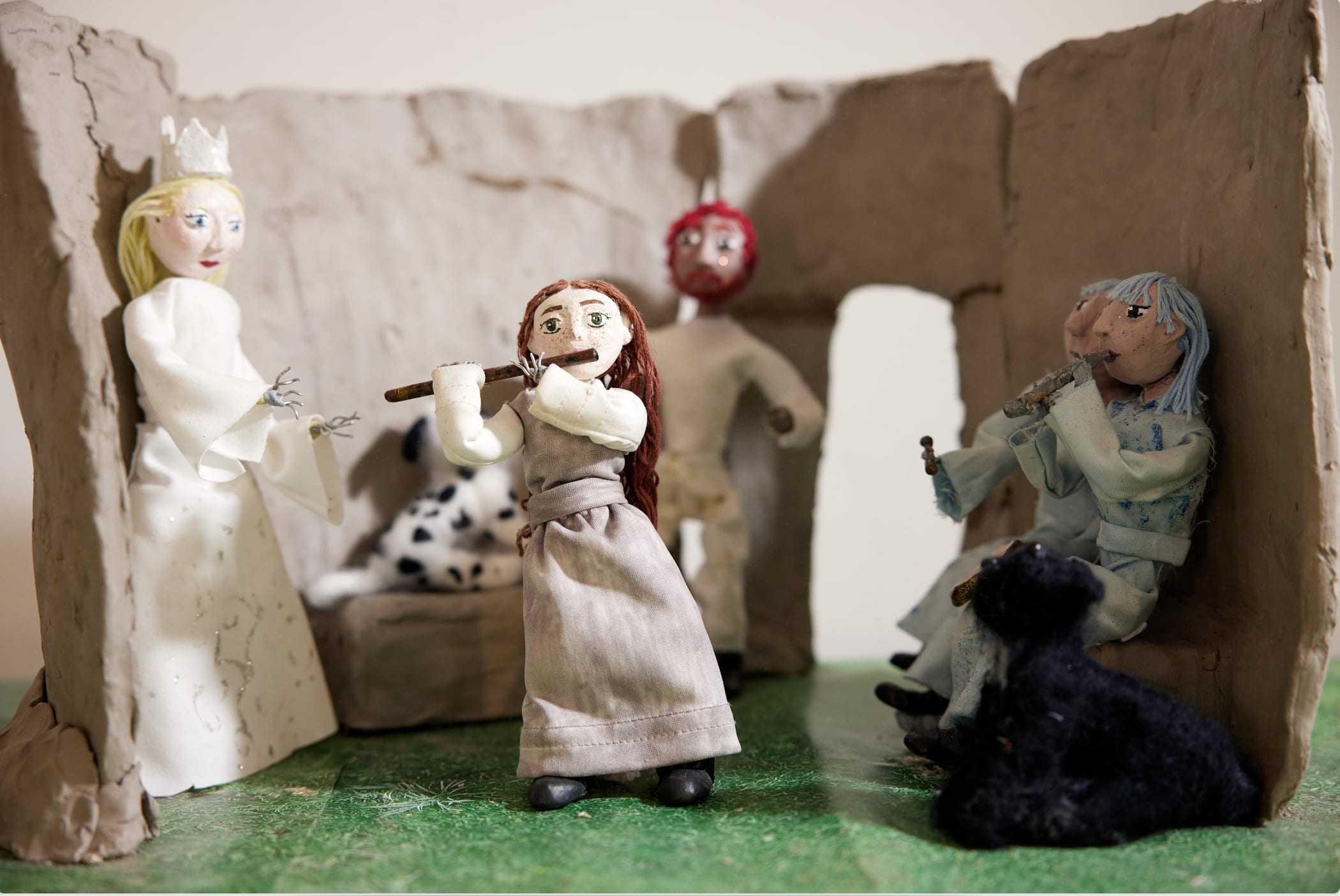 Small clay figures in a cave, three of them play woodwind instruments as one figure with a crown and a dog looks on
