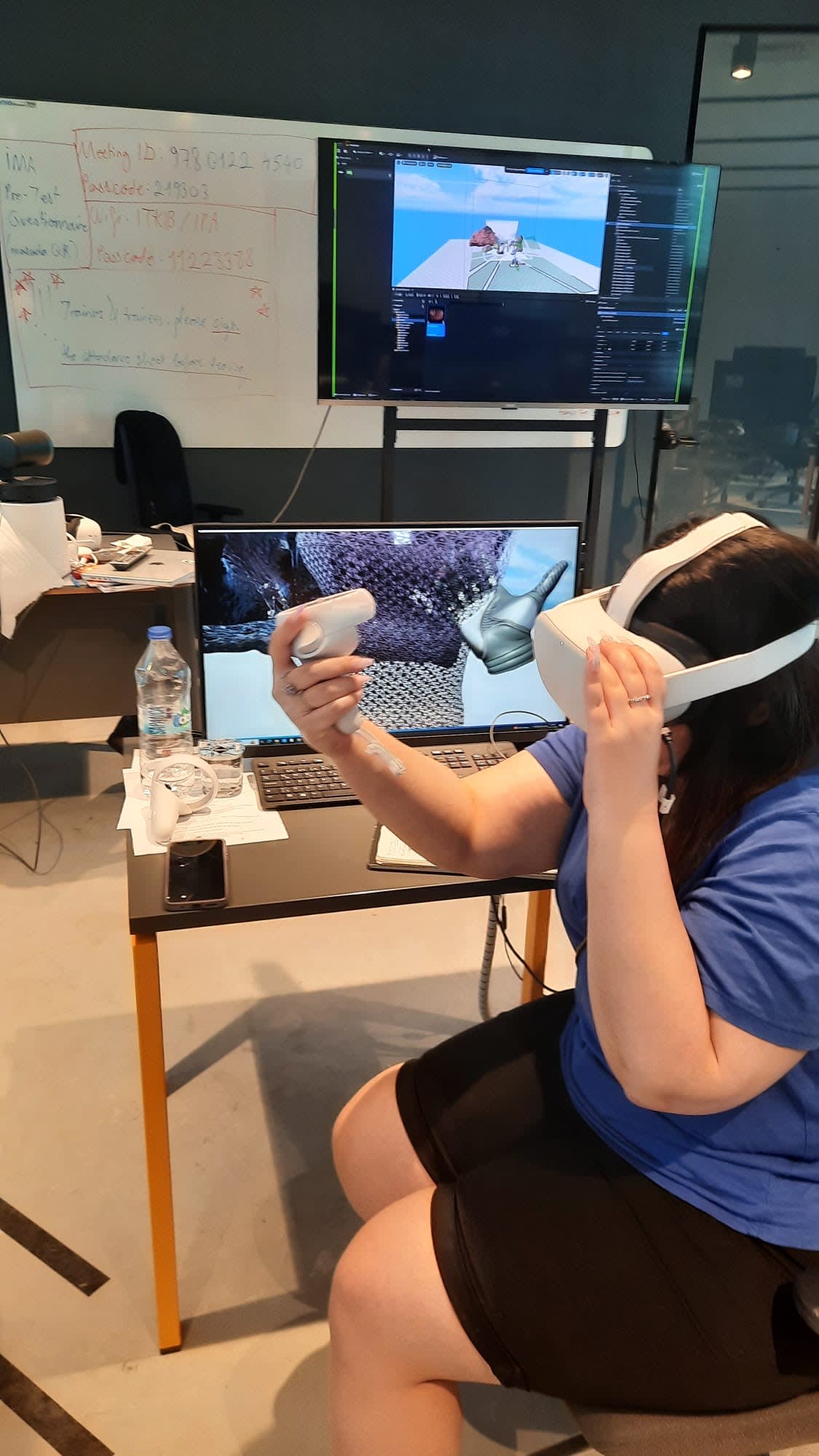A person sits in front of a screen with a 3D rendering on it while wearing a VR headset