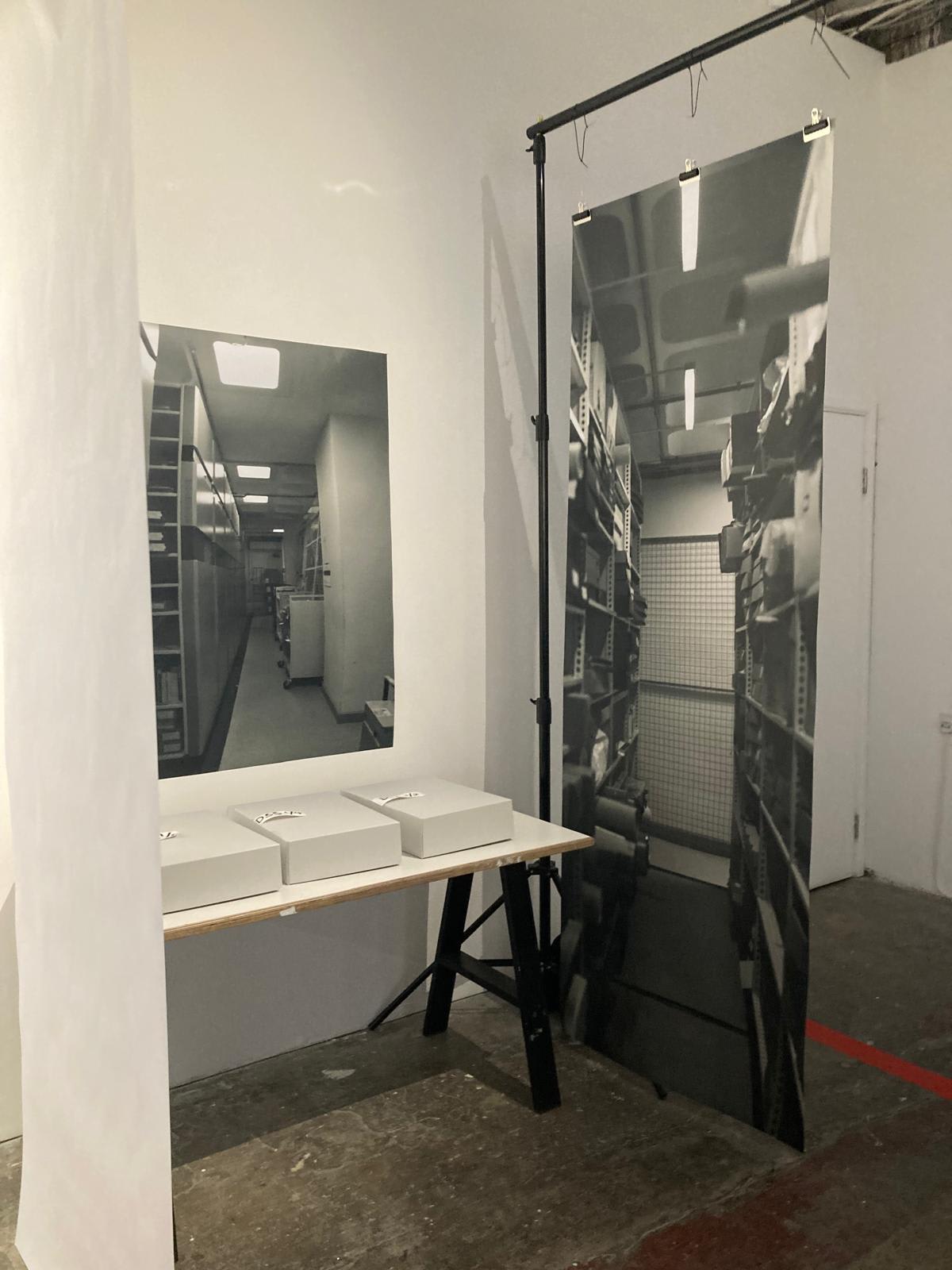 An installation with a table archive boxes and large format photographic prints mimicking the inside of an archive store.