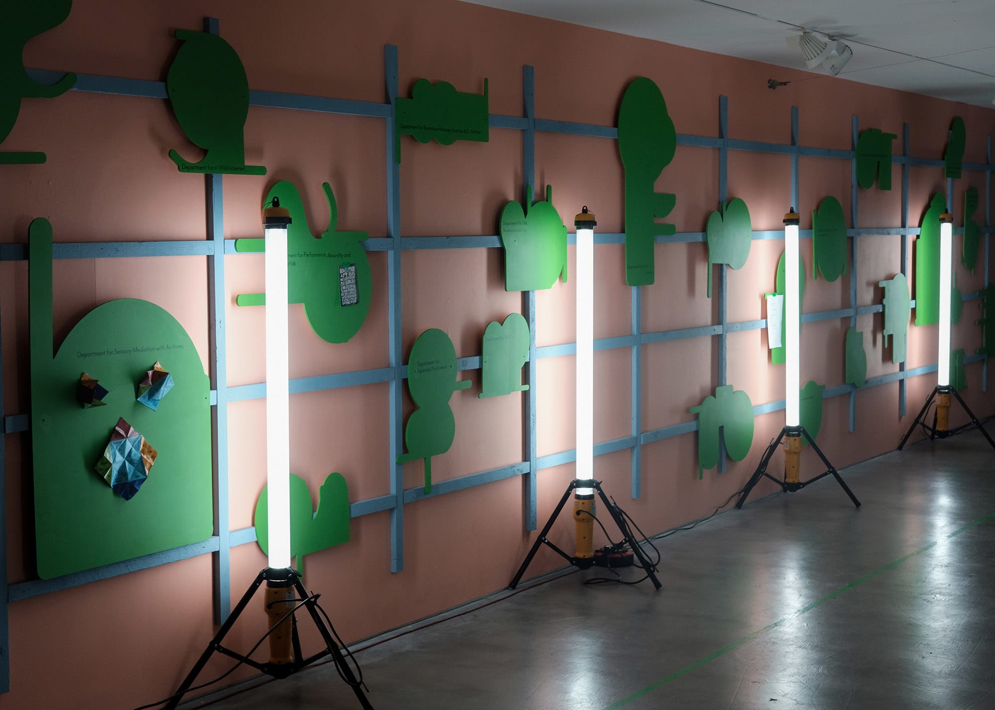 Green wooden shapes mounted on grid structure on a terracotta coloured wall in Camberwell Space gallery with free standing strip lights in front of it..