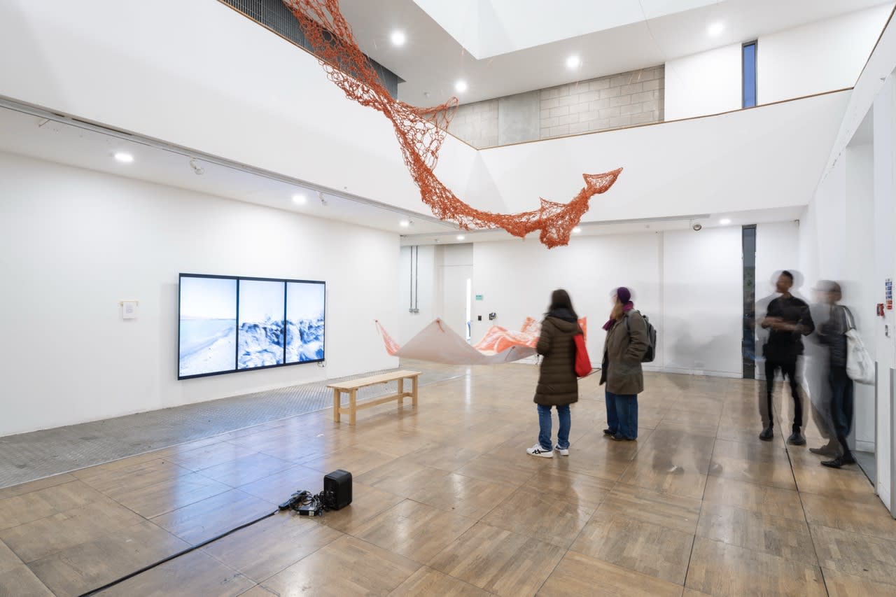 Exhibition photography showing a rope-like red structure hanging from a gallery ceiling. 