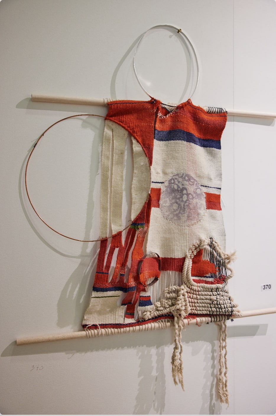 A red, white and blue tapestry made from dyed and torn canvas, string and rope, rope hung in between two wooden rods.