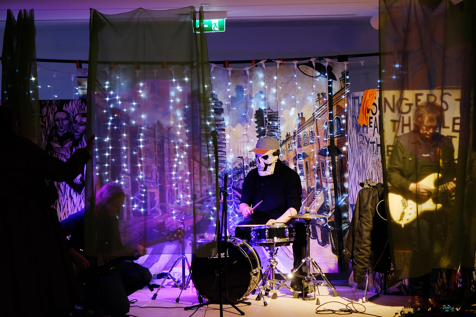Man playing drums wearing a skull mask and baseball cap surrounded by fairy lights.