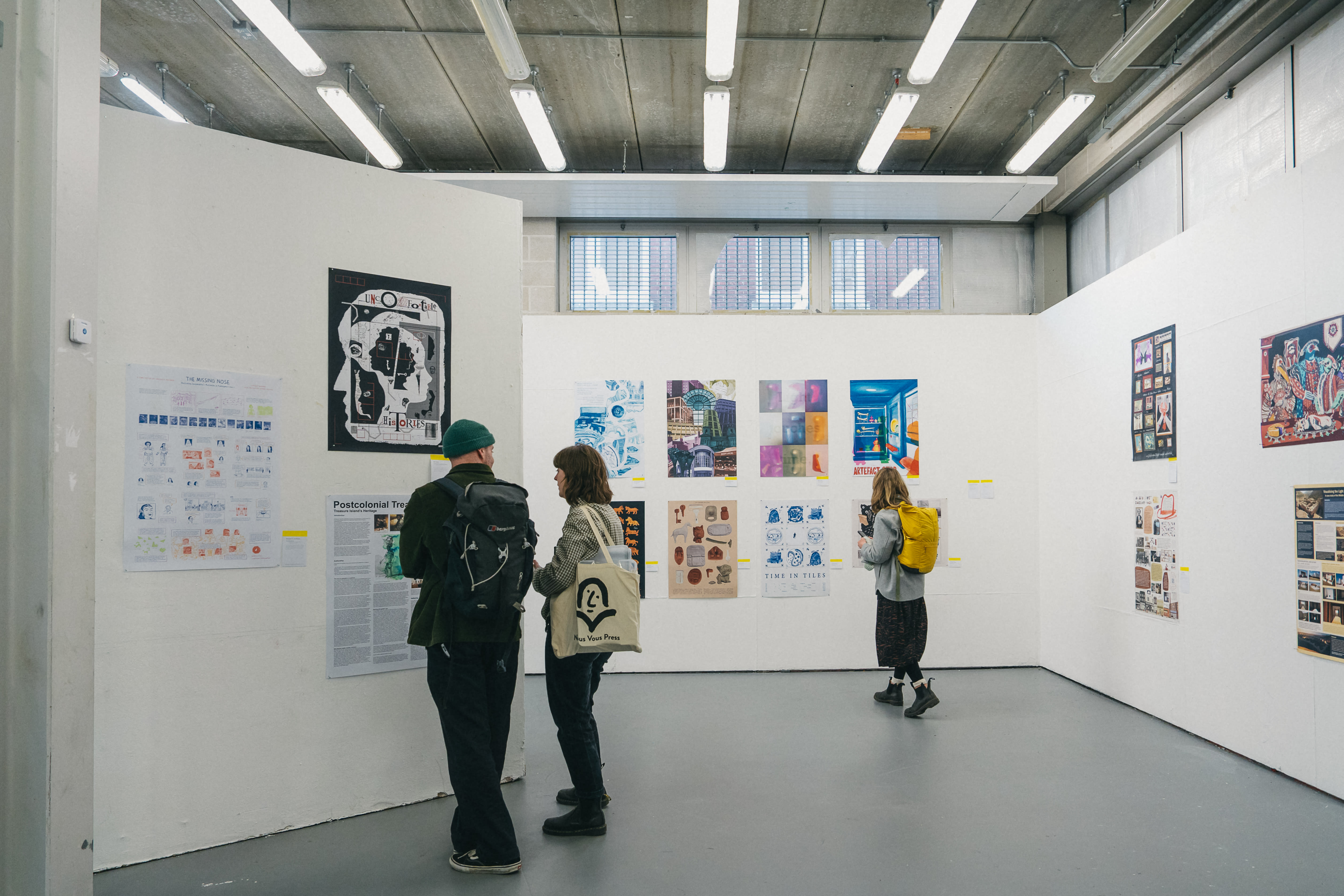 A gallery with white walls displays colorful artwork and posters. Three people view the art; one has a yellow backpack, while two converse. The setting feels modern and engaging.
