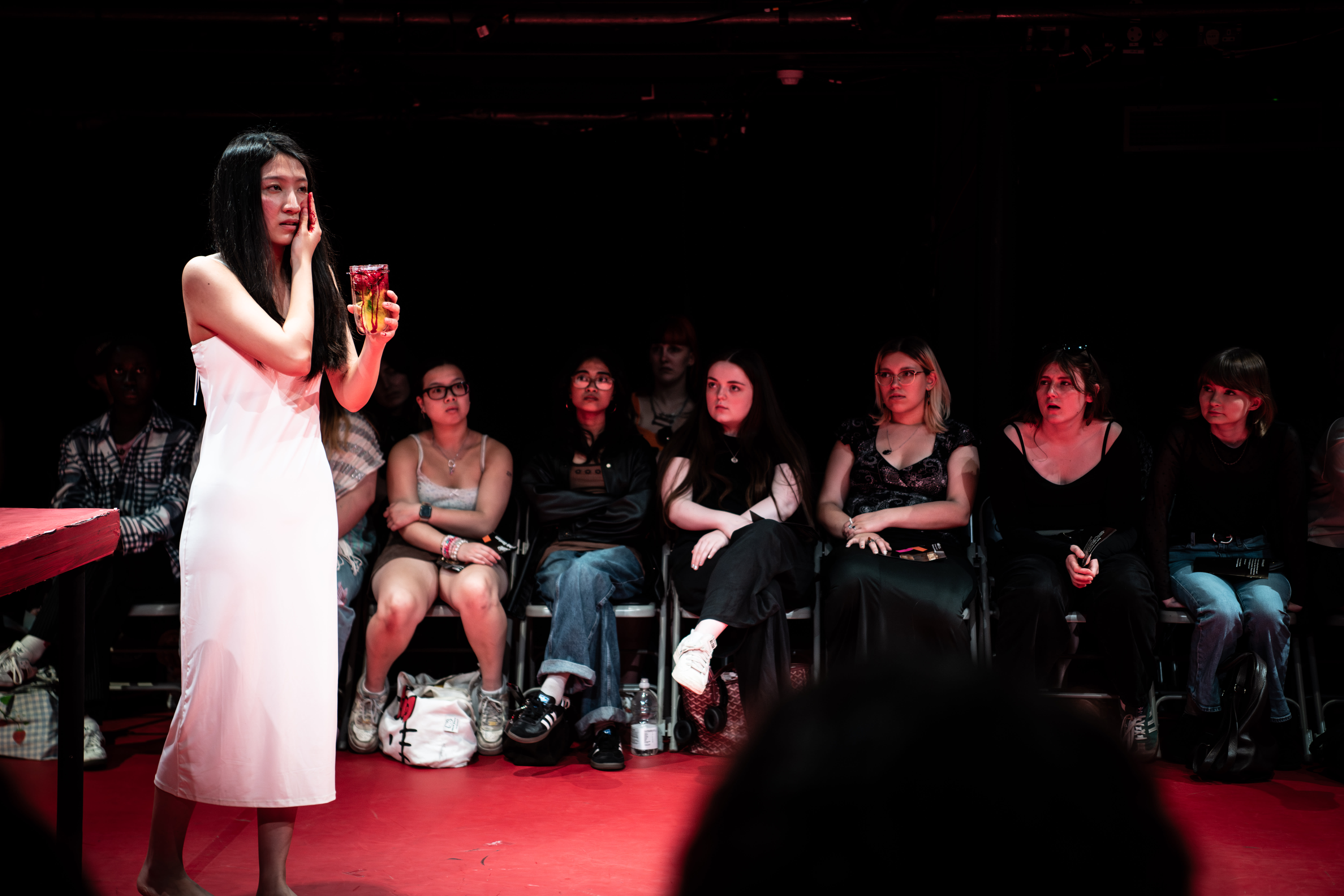 A performer in a white dress holds a drink, standing before an engaged audience seated in a dimly lit theater.