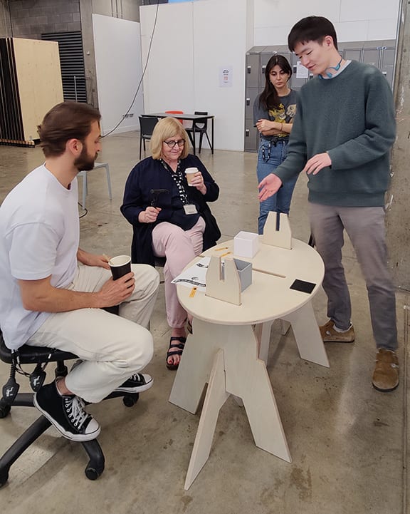 Group of people standing and sitting around a table examining a furniture prototype.