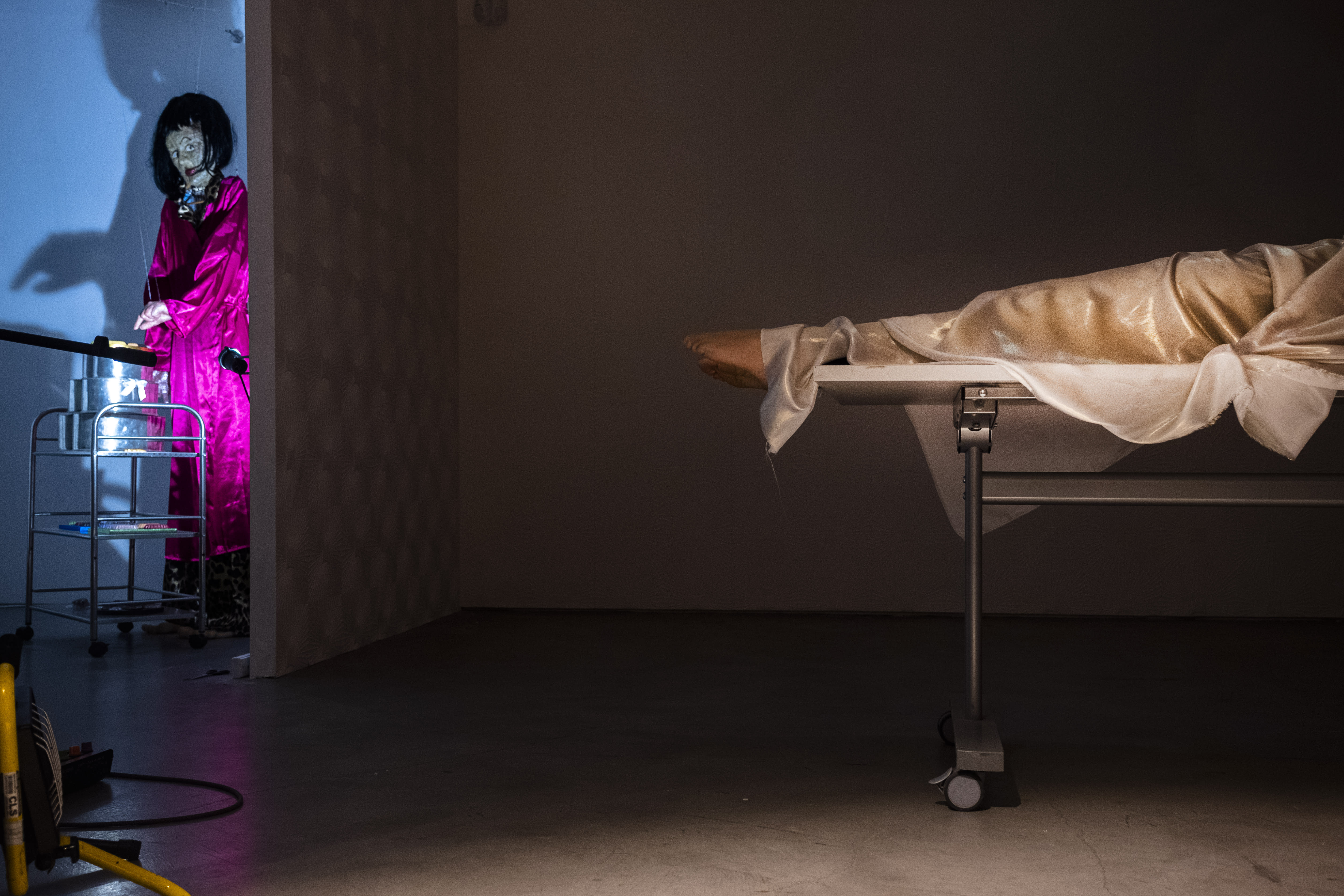 A doll in a pink satin robe looks around the corner to a body with their feet hanging off a table