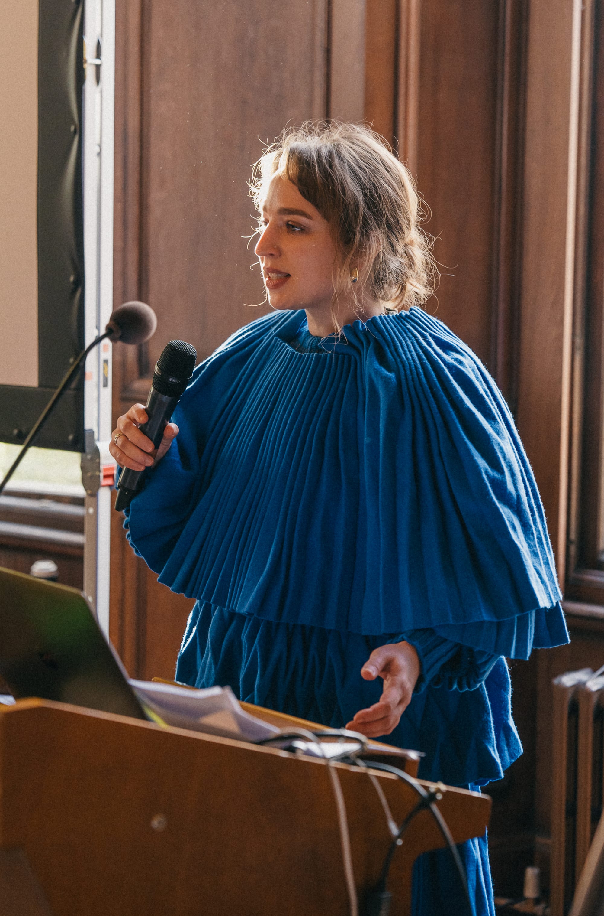 Woman in a blue pleated outfit speaking with a microphone.