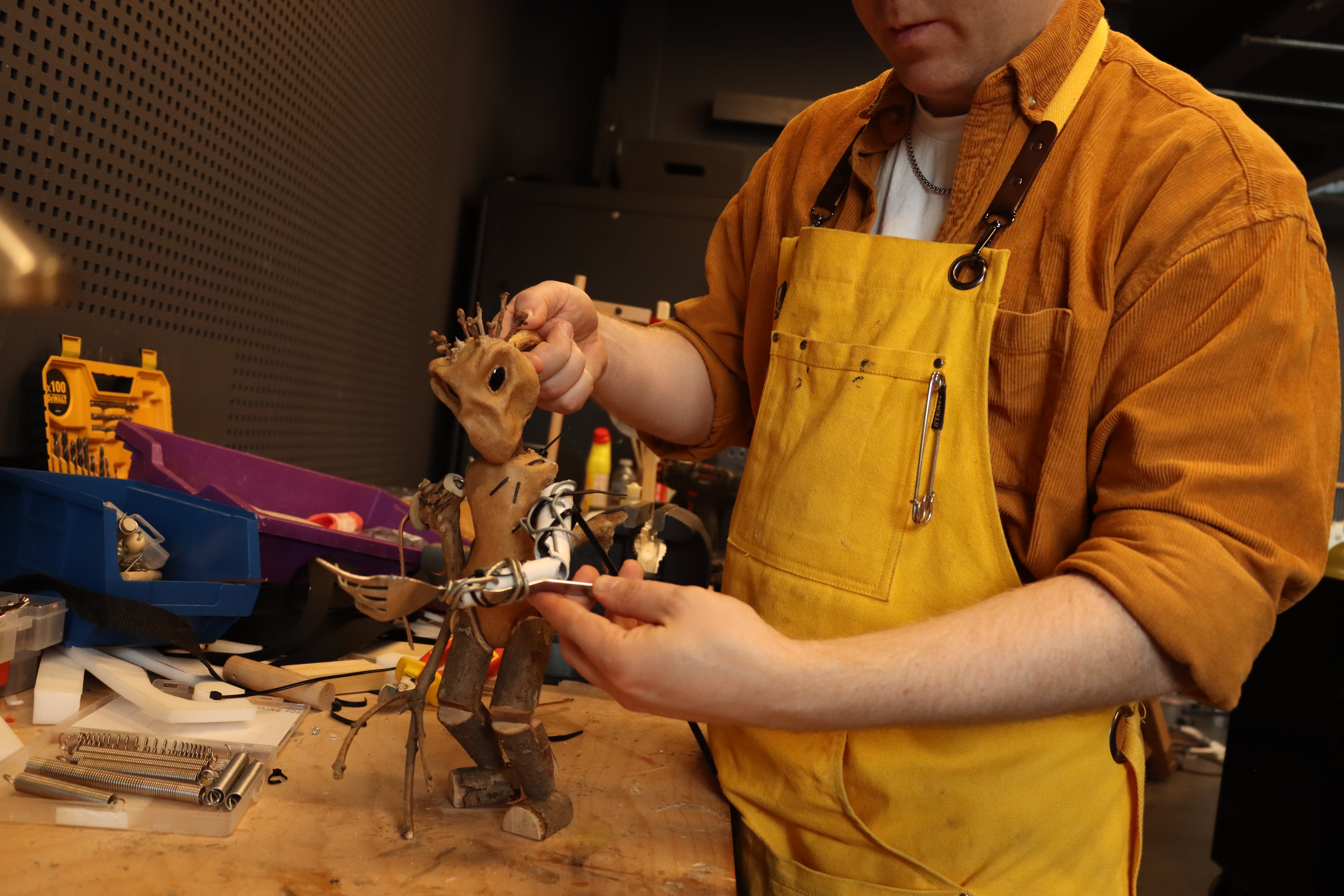 A person in a yellow apron intricately crafts a wooden figure on a workbench, surrounded by tools, conveying focus and creativity.
