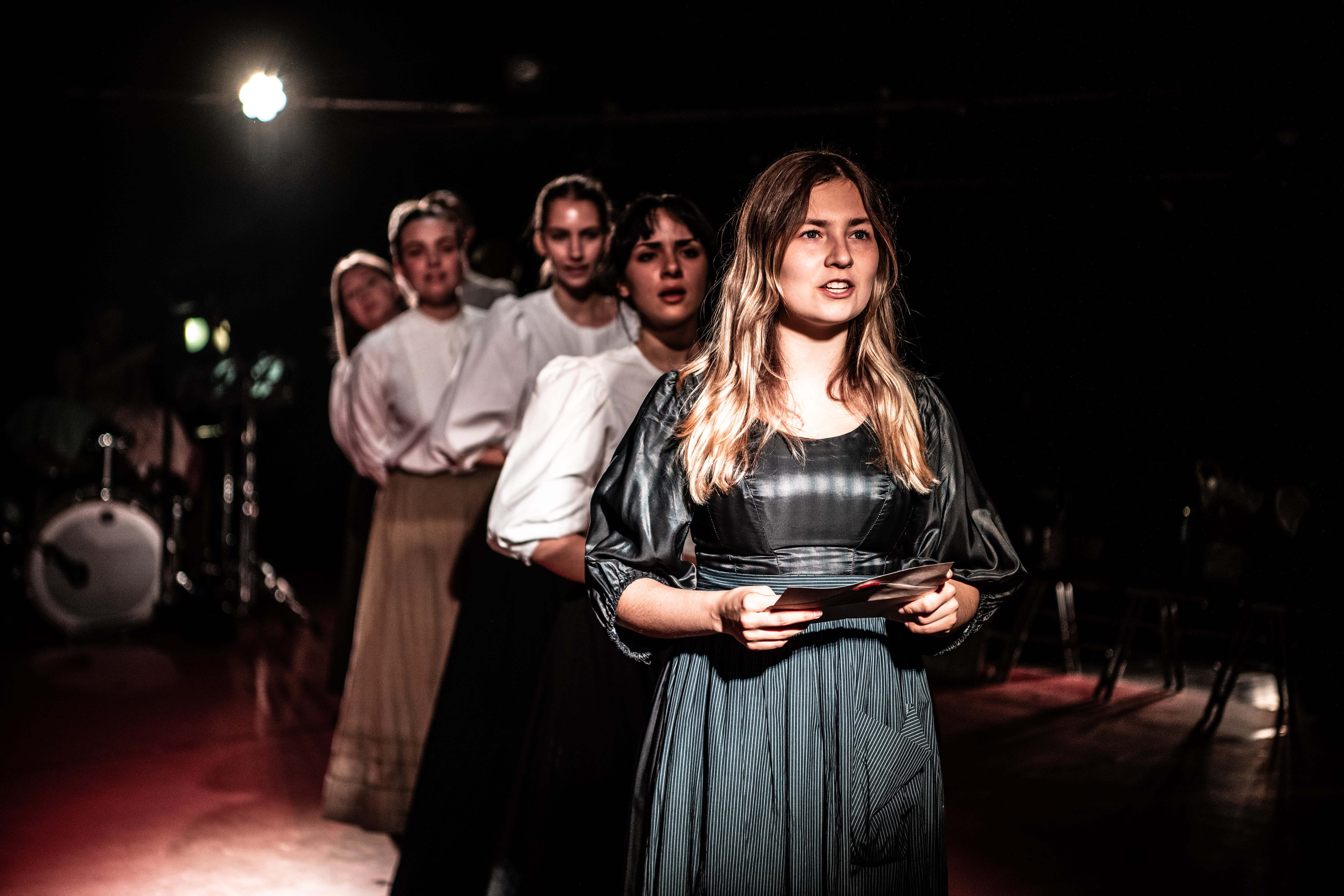 A group of women in period costumes stand in a line on stage, with the woman in front holding papers and speaking, under dramatic lighting.