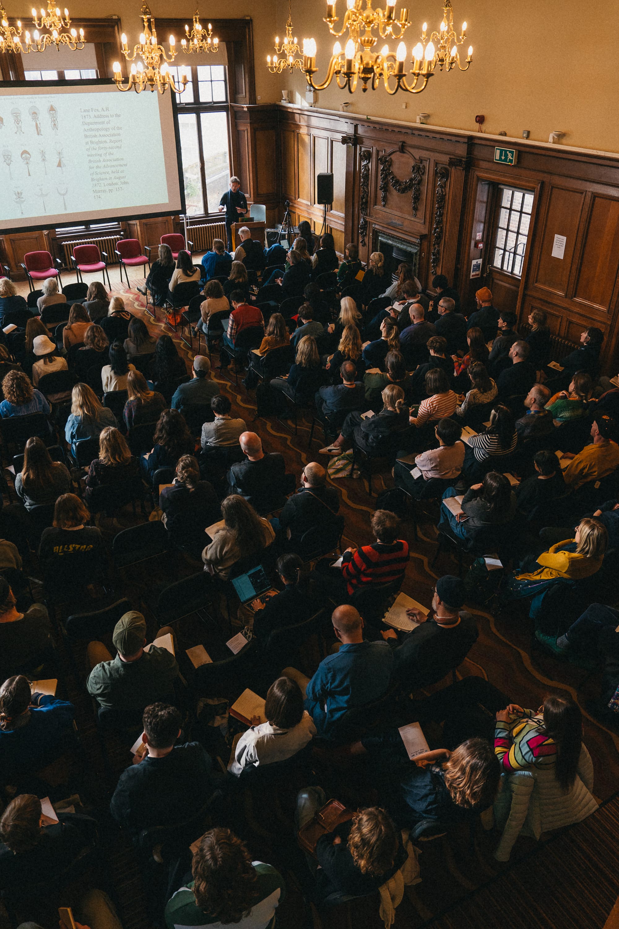 Aerial photo of a seated audience in front of a giant screen in a large wood panelled room with chandeliers.