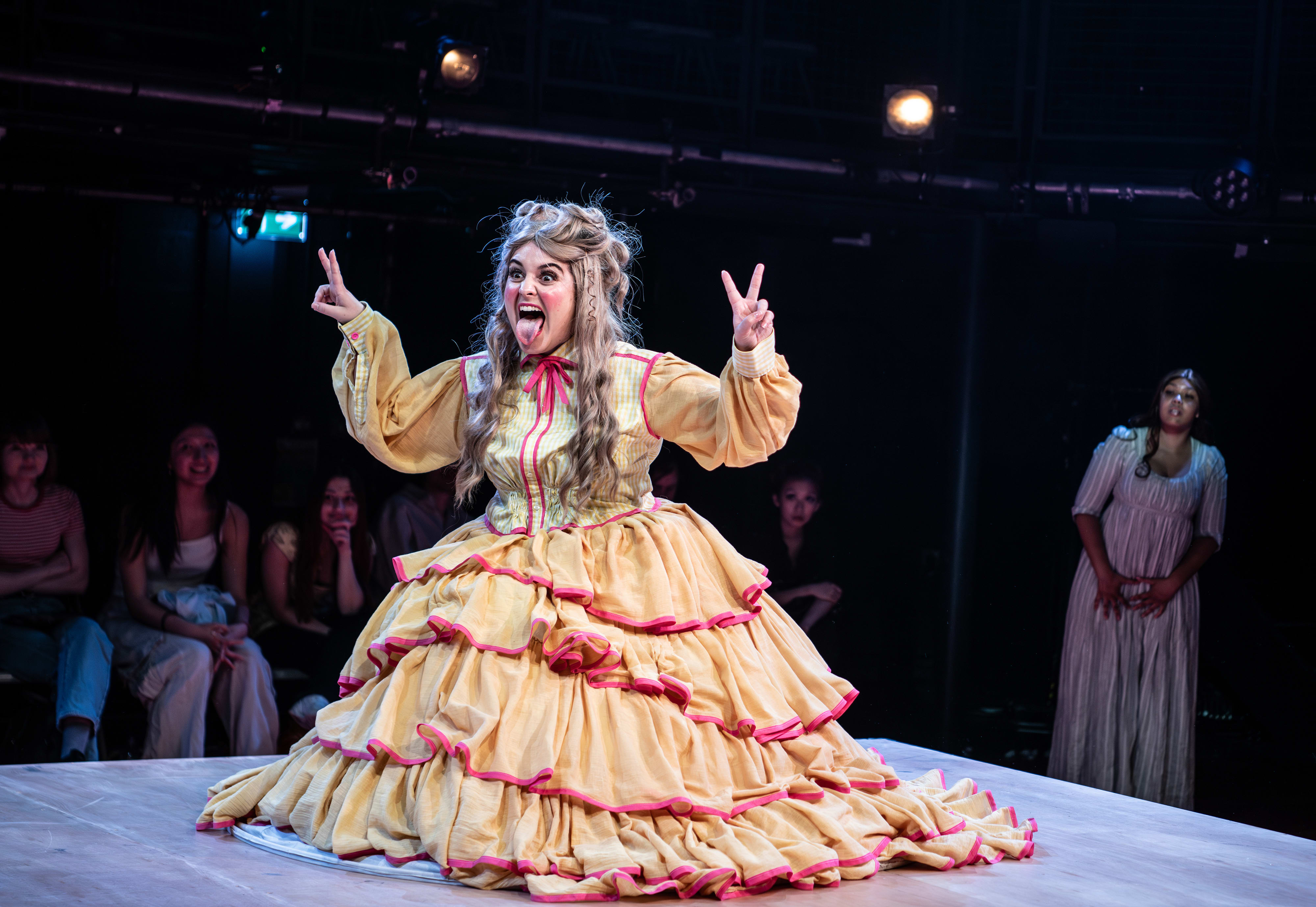A woman in a ruffled yellow dress kneels on stage, making peace signs and sticking out her tongue, while audience members watch in the background.