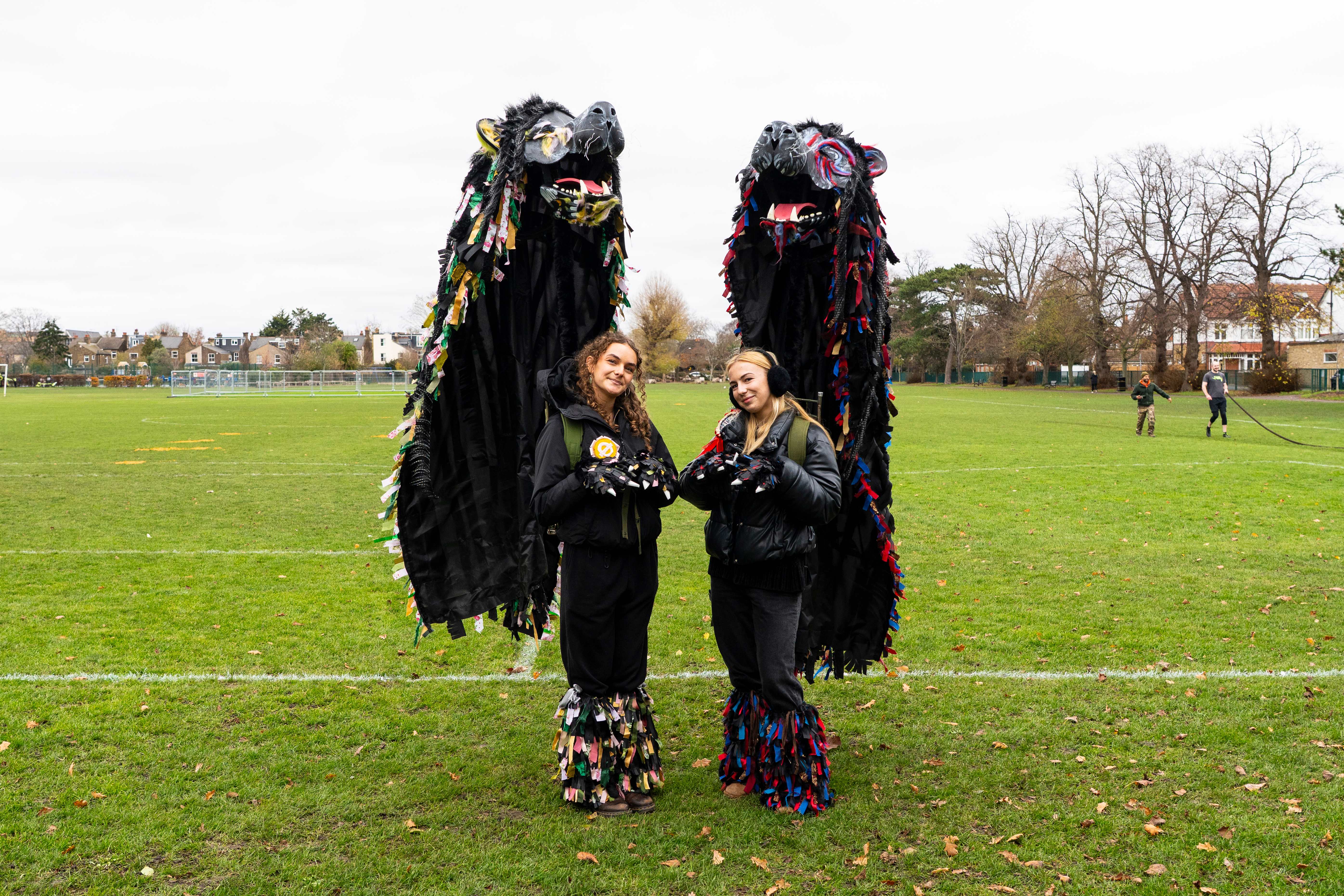 Two people stand on a grassy field, dressed in tall, colorful dragon costumes with large black capes. Both smile, creating a festive, joyful mood.