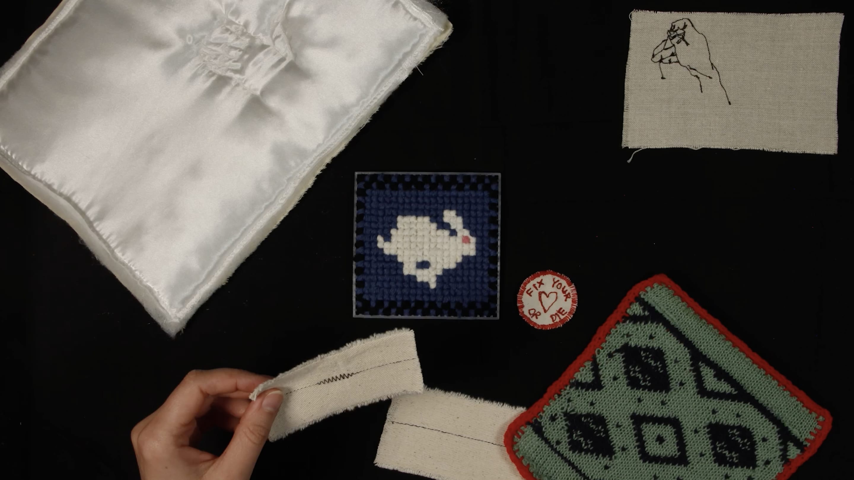 White, green and blue pieces of fabric placed on a black background with a hand holding a piece of fabric next to an embroidery of a rabbit.