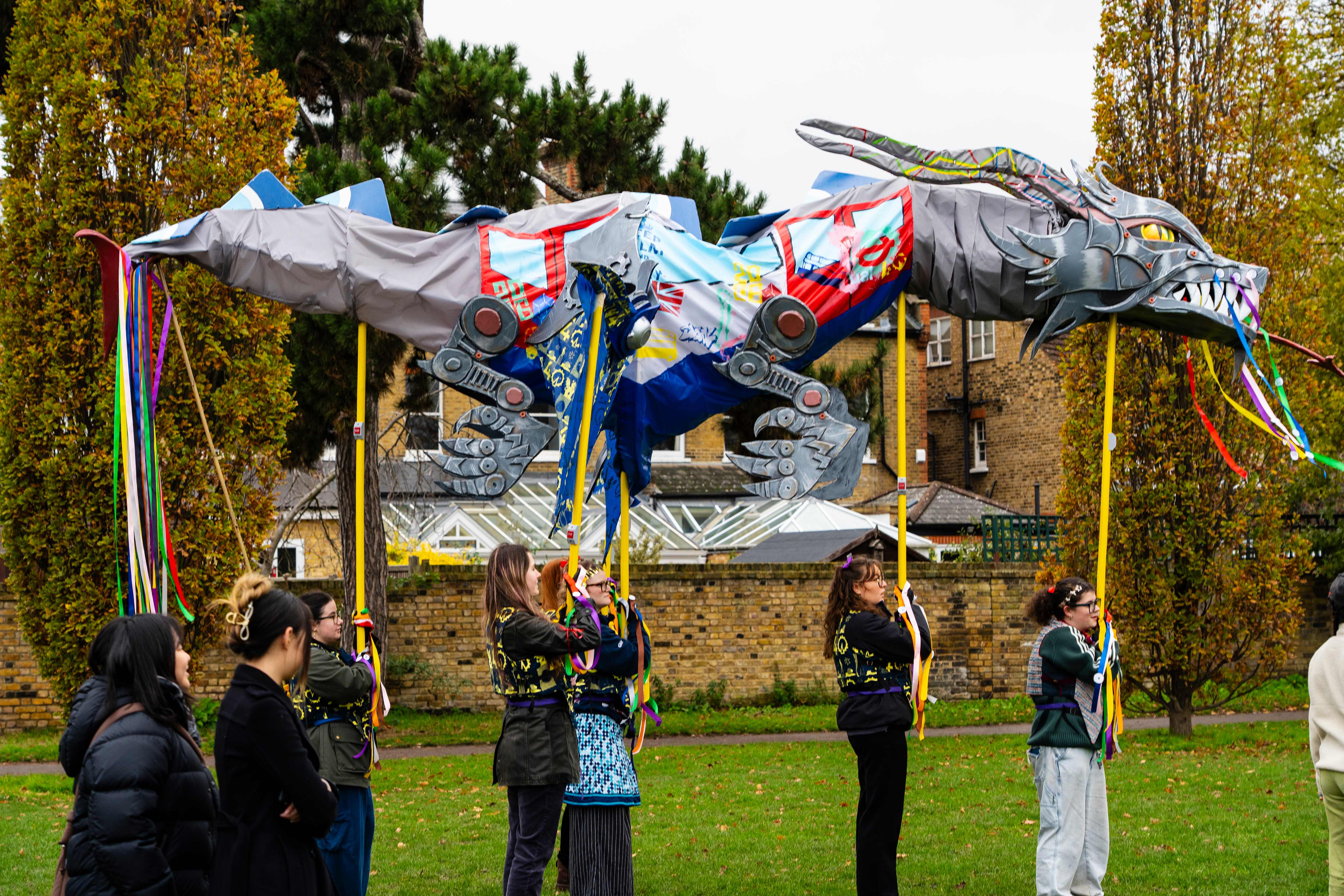People carry a colorful dragon puppet adorned with flags and ribbons in a park, set against a backdrop of trees and brick buildings.