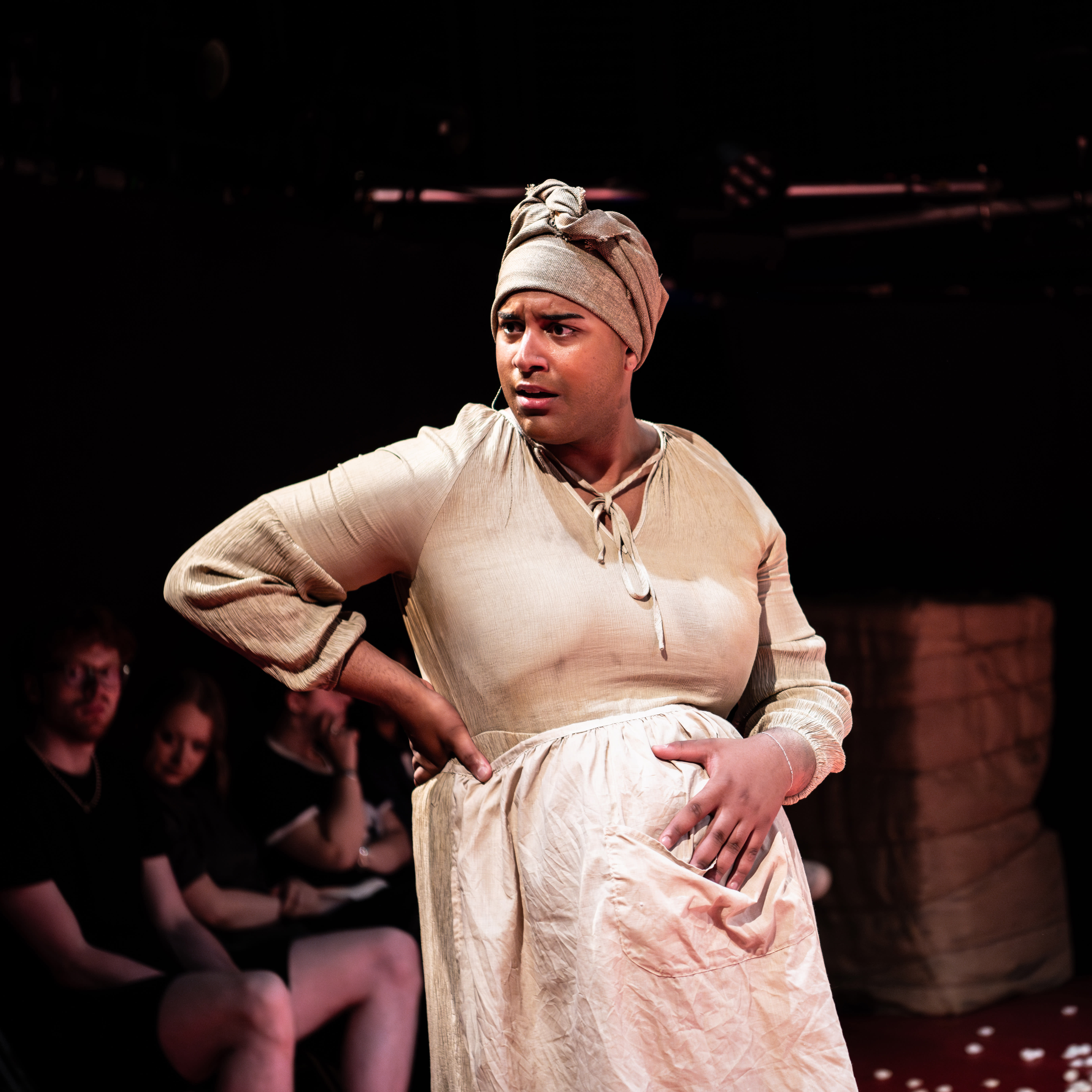 A performer in a light-colored dress and headscarf stands confidently with hands on hips, surrounded by an audience in a dimly lit theater.