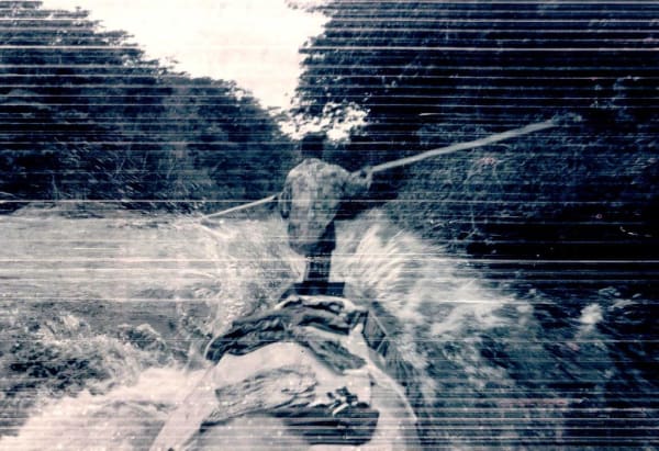 A grainy black and white image showing the back of a man steering a boat downriver
