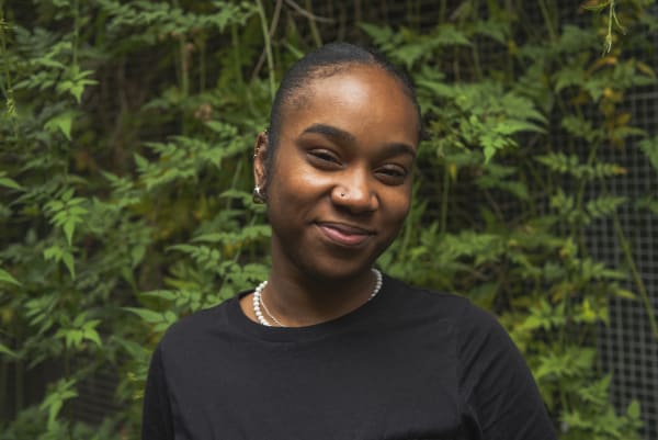 Photo of young woman in black top, smiling, standing infront of a background of green leaves.
