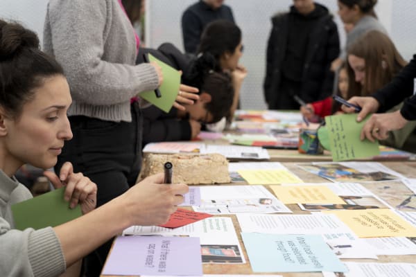 Close-up of a person kneeling to write something down on a piece of paper, while surrounded by others in a green house