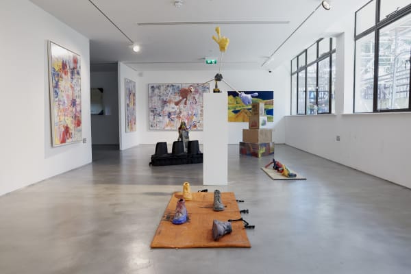 This image shows a contemporary art gallery space featuring various sculptures and abstract artworks. The sculptures include vibrant, unconventional forms displayed on the floor and pedestals, with painted canvases on the walls. The large windows to the right let in natural light, enhancing the clean, minimalist gallery atmosphere