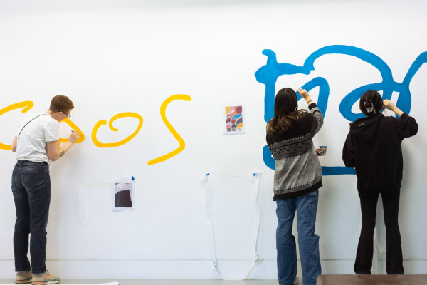 Students with their backs to camera painting large lettering in blue and yellow on a white wall