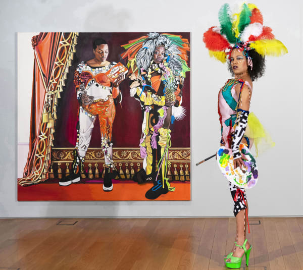 A person standing side-on in green heels looking at the camera in a colourful costume and headpiece. She has a painting palette in one hand and a paintbrush in the other. Behind her is a painting of two women. 