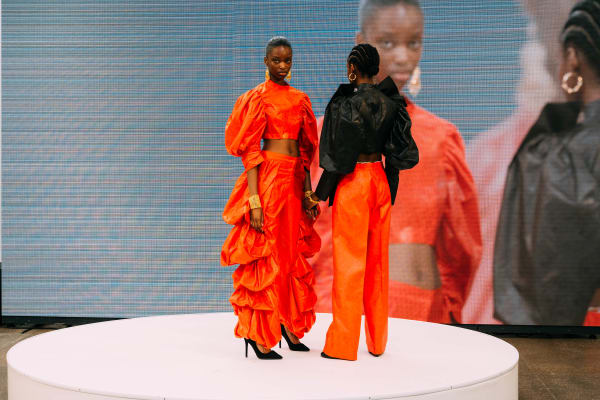 Two models on stage, left wearing a burnt-orange dress and the right wearing a black top with burnt orange pants