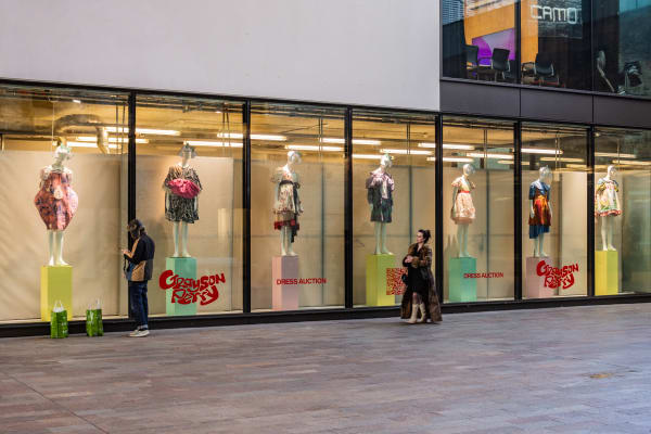 A wide shot of windows displaying an array of dresses on mannequins styled like Grayson Perry's Claire character. Red vinyl lettering says the words Grayson Perry + Dress Auction in creative type.