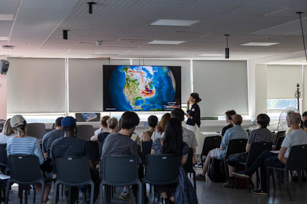 A group of students listen attentively to a presentation about design, with an image of the world projected on the screen.