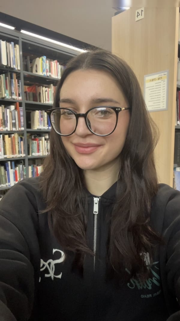 Headshot of the student Clara Popescu. She's shown with long dark hair and glasses smiles in a library, with bookshelves in the background. 