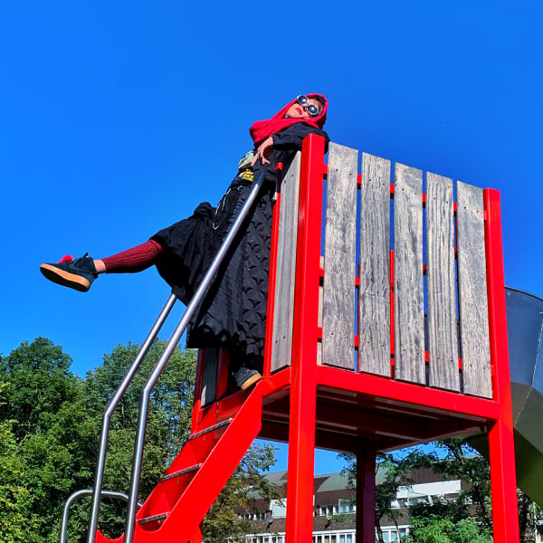 A woman poses in a red outfit on a red outdoor structure with her leg kicked out straight. The sky is clear and blue.