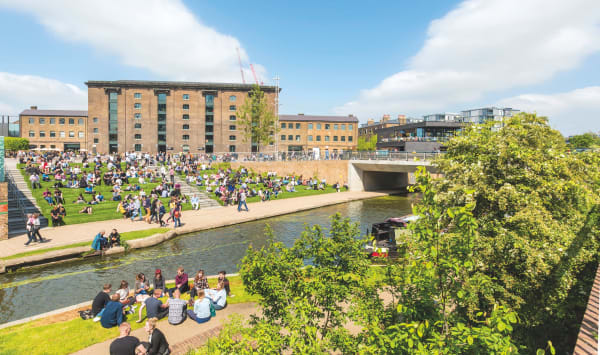 The image depicts an exterior view of the Granary Building, showing a vibrant outdoor scene with many people enjoying the weather on the green lawn areas and along the canal. The setting features large, historic brick buildings in the background, while the foreground includes people sitting on steps and by the water's edge, socializing and relaxing. The overall atmosphere is lively and communal, with clear blue skies enhancing the inviting environment.
