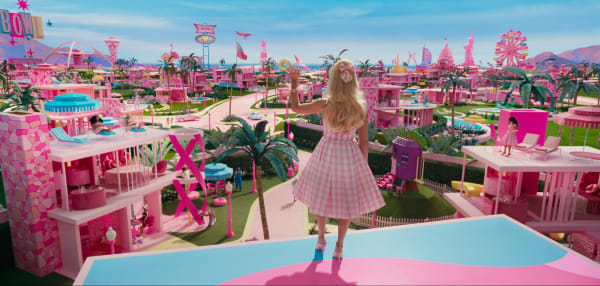 Margot Robbie stands on top of a blue and pink building and is waving across Barbie Land: a fictional land where most buildings are pink, with palm trees and a lush blue skyline in the background.
