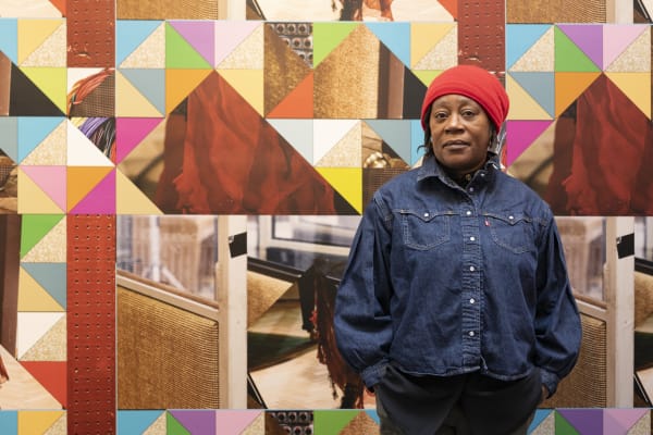 Sonia Boyce standing in front of artwork. Behind her is a colourful tapestry of geometric shapes. 