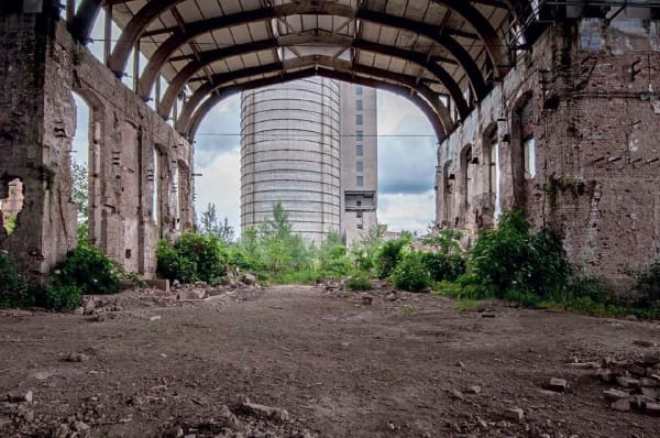 A derelict industrial building with an internal archway overgrown with grass and shrubs and a newer brutalist style building beyond the arch