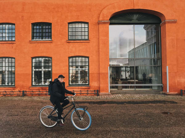 A bright orange building in the background with a young Asian man on a bike cycling past it