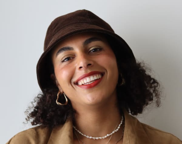 A person wearing a bucket hat and smiling with their chin tilted up. They are standing against a white wall.